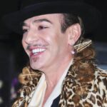 John Galliano z szerokim uśmiechem. Artysta ma na sobie czarny kapelusz i lekki płaszcz w panterkę.