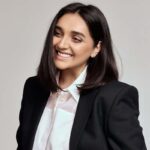Elvira Gasanova – projektantka, która ubiera Zełenskiego. Kim ona jest? Zdjęcie uśmiechniętej projektantki pozuje w eleganckiej czarnej marynarce i białej koszuli na jasnym tle.