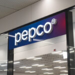 Szyld sklepu Pepco, należącego do Pepco Group, który właśnie sprzedał sieć dyskontów Poundland. Szyld jest niebiesko-fioletowy, z płynnym przejściem kolorów. Napisy są białe. Na zdjęciu widać również sufit sklepu z lampami.