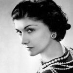 Coco Chanel na starej fotografii. Jej włosy ułożone są w fale retro, ma na sobie czarną sukienkę i sznur pereł. Siedzi tyłem do obiektywu, ale odwraca się, by spojrzeć prosto na widza.