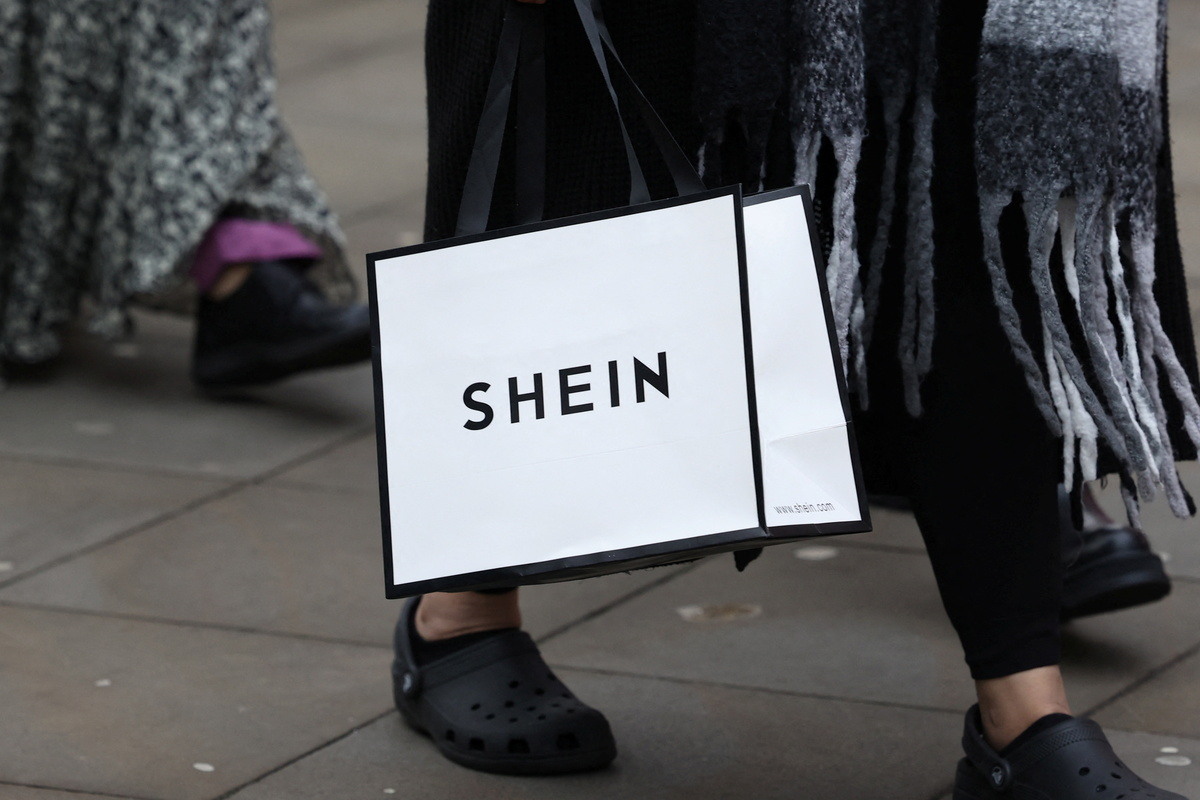 Chiński potentat ultra fast fashion – Shein – wchodzi na rynek handlu tradycyjnego. Już 5 listopada firma uruchomi swój pierwszy na świecie sklep stacjonarny z prawdziwego zdarzenia w paryskim domu towarowym BHV. Ta decyzja generuje spore oburzenie – od oporu władz Paryża, poprzez stowarzyszenia branżowe, aż po manifestacje samych pracowników BHV. Shein sklep stacjonarny - gdzie i kiedy otwarcie? Zbliżenie na białą torbę zakupową z czarnym napisem Shein. Torba jest niesiona przez kobietę w czarnych spodniach i czarnych butach typu Crocs. Widać długi szalik z frędzlami.