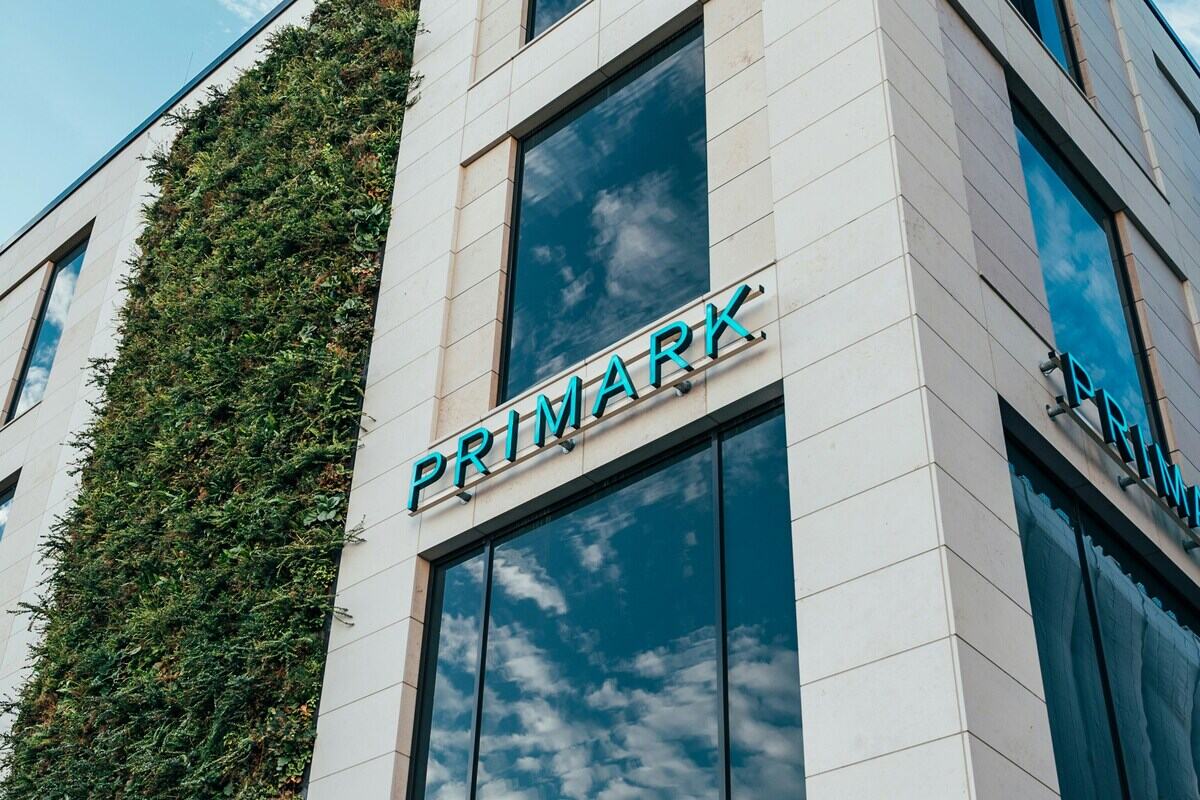 Zamknięcie sklepu Primark. Zewnętrzna fasada nowoczesnego, beżowego budynku, na którym widnieje neonowy, turkusowy napis Primark. Po lewej stronie elewacji znajduje się duża, pionowa zielona ściana z roślinności. W ciemnych oknach odbija się niebo z białymi chmurami.