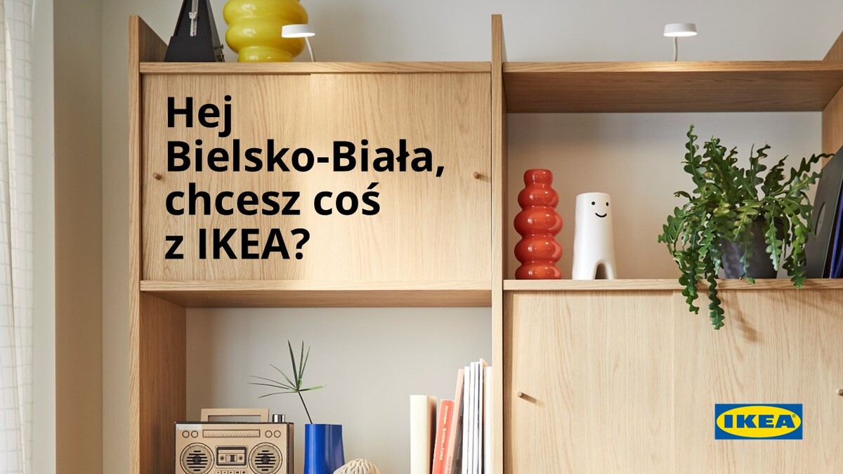 Zdjęcie regału wykonanego z jasnego drewna, na którym znajdują się książki i elementy dekoracyjne, takie jak czerwony wazon w kształcie kuli oraz biała figurka z uśmiechem. Na zdjęciu umieszczony jest czarny napis: &quot;Hej Bielsko-Biała, chcesz coś z IKEA?&quot;, a w prawym dolnym rogu widnieje logo IKEA. Jest to materiał reklamowy.