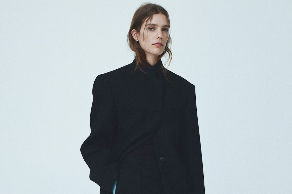 Wyniki finansowe H&amp;M Q3 2025. Fotografia modelki w strukturalnej, powiększonej czarnej marynarce z wyraźnie zaakcentowanymi, rozłożystymi ramionami oraz czarnym golfem (lub wysokim kołnierzem) pod spodem. Dama ma swobodnie upięte włosy i spogląda wprost w kamerę. Tło jest błękitne.