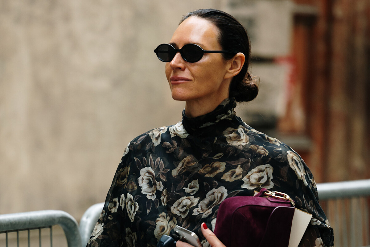 Ningi Research atakuje CCC. Portret stylowej damy w okularach słonecznych, prawdopodobnie sfotografowanej na ulicy podczas jakiegoś wydarzenia ze świata mody (być może Paris Haute Couture, jak sugeruje nazwa pliku). Ma ciemne, idealnie uczesane włosy związane w kok z tyłu głowy. Ma na sobie czarną bluzkę lub kreację z wysokim dekoltem, przyozdobioną wzorem w jasne (białe lub kremowe) róże oraz listowie. Okulary przeciwsłoneczne są niewielkie, owalne i posiadają czarne ramki. W lewej dłoni kobieta ściska smartfon i ewentualnie inne drobiazgi, a w prawej ręce ciemnobordową zamszową torebkę lub puzderko oraz jasną książkę/notes. W oddali widoczny jest zamazany fragment szarej ściany budynku oraz metalowa barierka. Spogląda w lewo poza obszar zdjęcia.