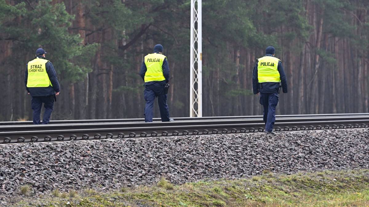 Trzech policjant&oacute;w w ż&oacute;łtych kamizelkach odblaskowych stoi na torach kolejowych, patrząc wzdłuż nich.