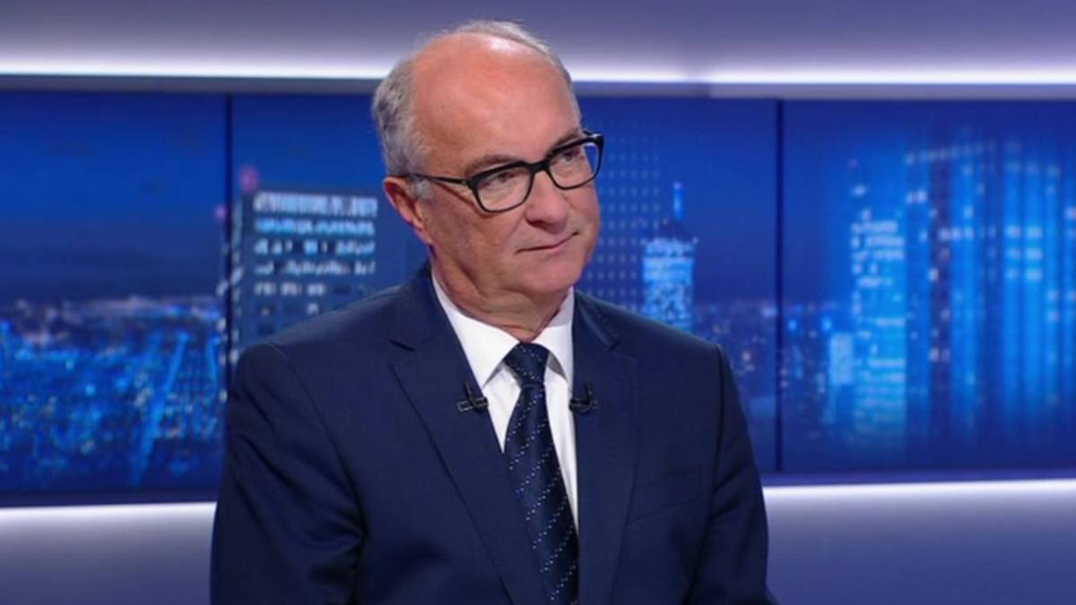 Mężczyzna w średnim wieku w granatowym garniturze, białej koszuli i ciemnym krawacie, noszący okulary w czarnych oprawkach, siedzi w studiu telewizyjnym. Za nim znajduje się ekran z widokiem nocnej panoramy miasta z oświetlonymi budynkami.