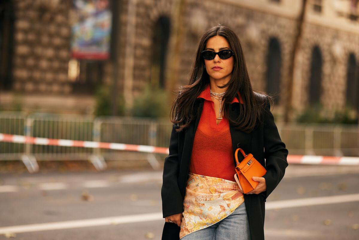 Bloomberg informuje, że rosyjskie przedsiębiorstwa składają podania o rejestrację brandów pozostawionych przez zagraniczne korporacje w roku 2022. Od początku bieżącego roku dotyczy to ponad trzystu spółek, w tym między innymi Victoria’s Secret, Ericsson, Inditex i Nokia. Trzyletni okres braku aktywności stał się bodźcem do składania wniosków przez miejscowe jednostki, które usiłują zagarnąć nieużywane, lecz rozpoznawalne szyldy. Przejęcia marek, znaków towarowych Rosja. Na zdjęciu: Kobieta podczas fashion weeku przemierzająca ulicę w czarnym okryciu, jaskrawopomarańczowym pulowerze i jedwabnej chuście zawiązanej w talii, z niewielką pomarańczową torebką w ręce i szkłami przeciwsłonecznymi.