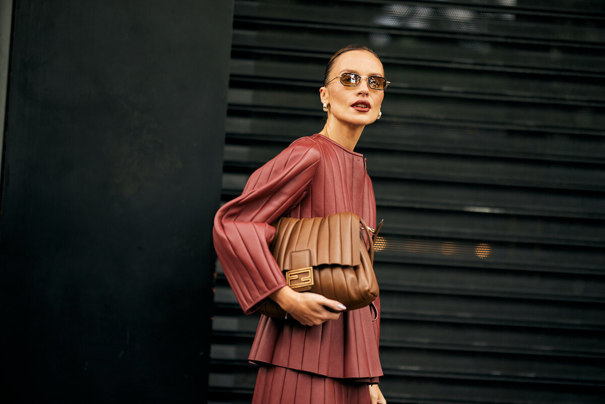 Kobieta w bordowym, pikowanym zestawie i z dużą brązową torebką na ramieniu stoi na tle ciemnej, industrialnej ściany. Stylizacja inspirowana modą premium symbolizuje współczesne najsilniejsze marki e-commerce 2025, które łączą estetykę, wiarygodność i rozpoznawalność w komunikacji.