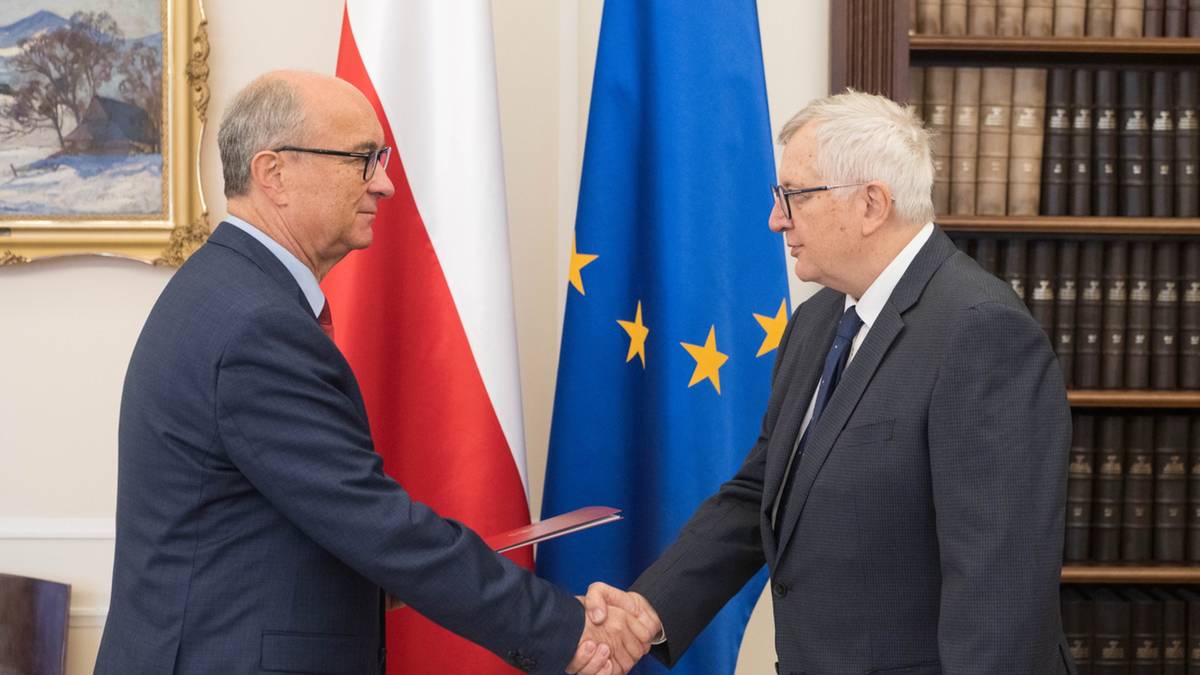 Dw&oacute;ch mężczyzn w garniturach ściska sobie dłonie przed polską flagą i flagą Unii Europejskiej.