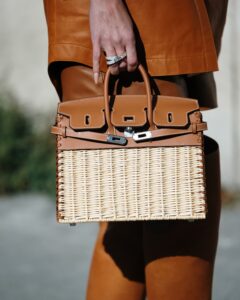Wyjątkowa, pleciona torba Hermès Kelly Picnic w barwie naturalnej rafii z brązowymi detalami skórzanymi i okuciami w kolorze srebra, trzymana przez kobietę w stylizacji z karmelowymi elementami odzieży skórzanej i botkami. Detal na dłoń z pierścionkiem.