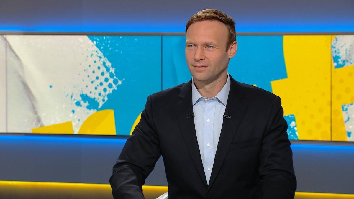 Mężczyzna w garniturze siedzi przed kolorowym tłem studia telewizyjnego.