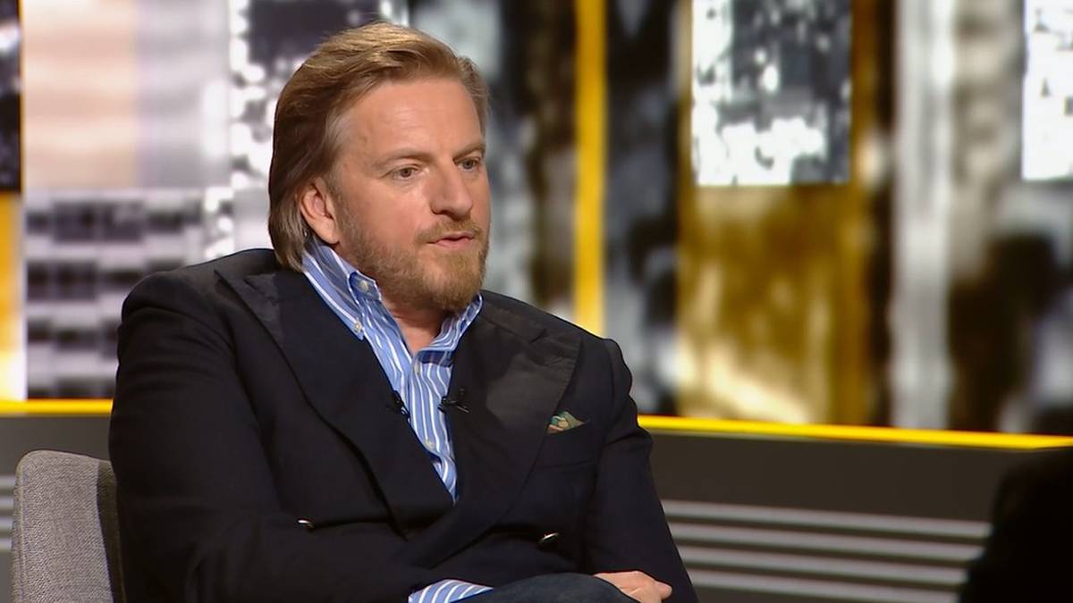 Mężczyzna z jasnymi włosami i brodą, ubrany w niebieską koszulę w paski i marynarkę, siedzi w fotelu.