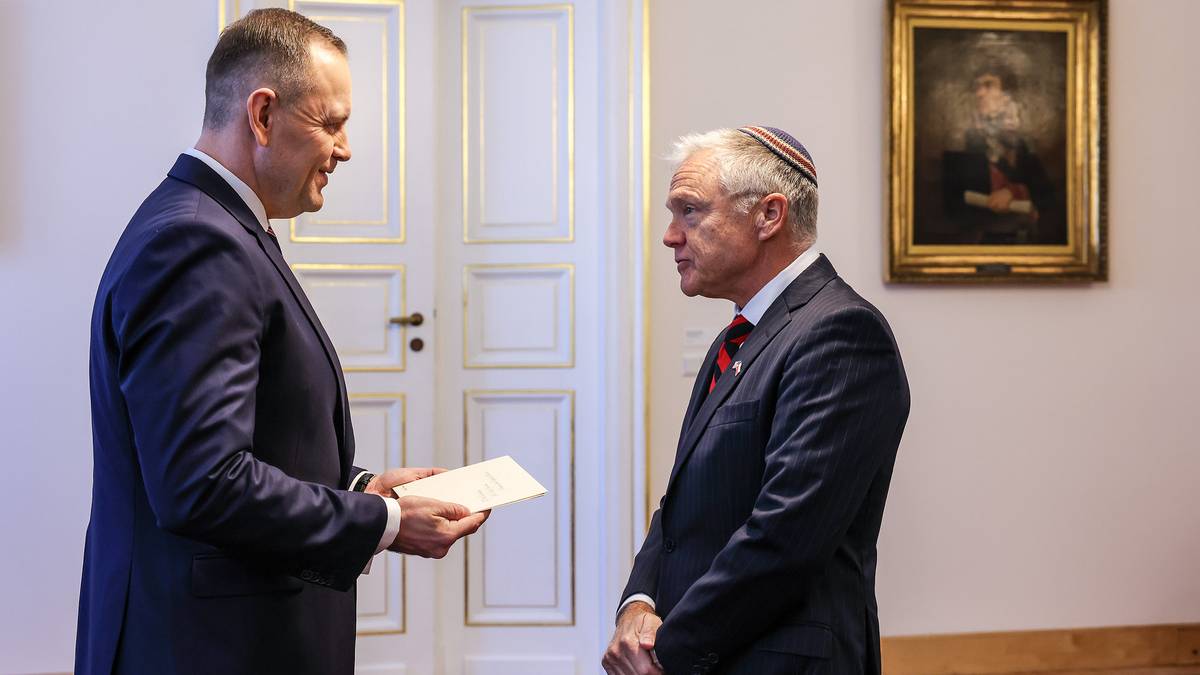 Dw&oacute;ch mężczyzn w eleganckich garniturach stoi naprzeciwko siebie w pomieszczeniu. Jeden z nich, po lewej, ubrany jest w ciemny garnitur i trzyma w ręku kartkę papieru, kt&oacute;rą podaje drugiemu mężczyźnie. Drugi mężczyzna, po prawej, ma na sobie ciemny garnitur z czerwoną poszetką i okrągłe okulary. Na ścianie w tle wisi obraz w złotej ramie. Scena sugeruje formalne spotkanie lub wręczenie dokumentu.