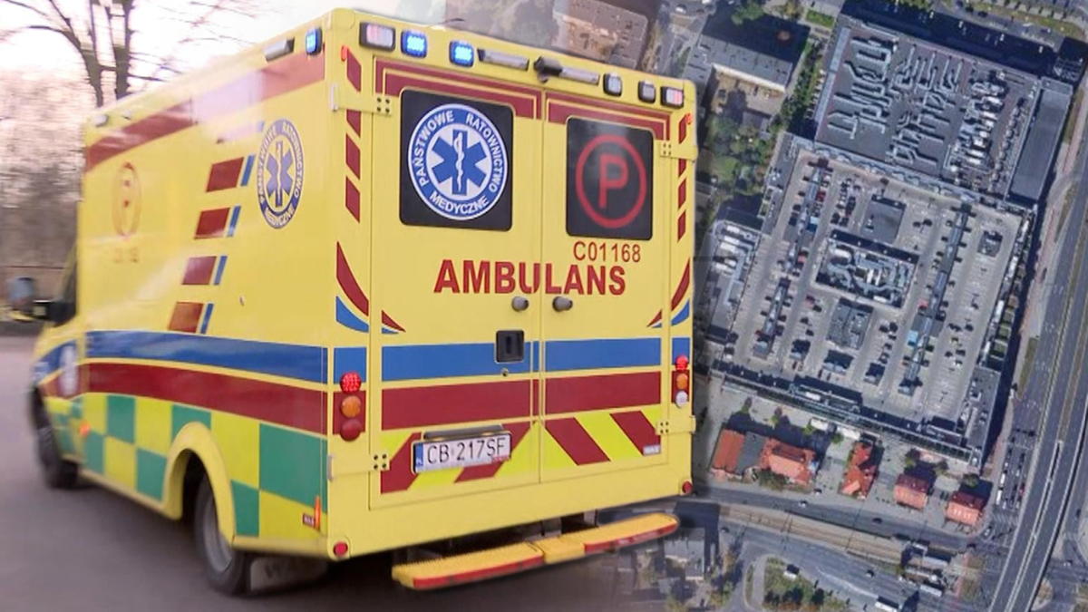 Karetka pogotowia z napisem &quot;AMBULANS&quot; na tle widoku z lotu ptaka na parking galerii handlowej.