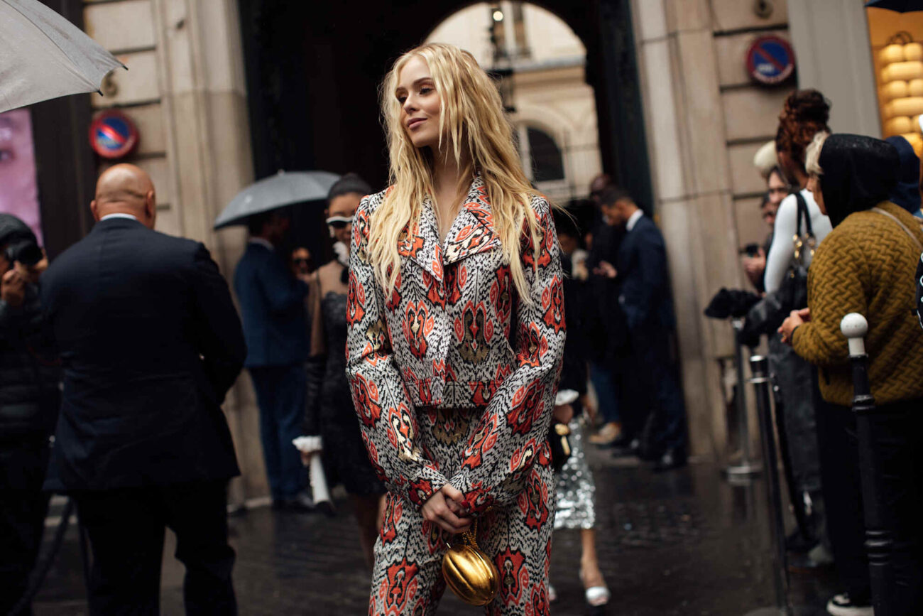 UE, cła na tanie paczki z Chin. Na zdjęciu widać kobietę ubraną w elegancki, deseńowy kostium w tonacjach czerwieni i beżu, stojącą na ulicy w otoczeniu innych uczestników wydarzenia związanego z modą. Scena najpewniej pochodzi z fashion week w jednym z europejskich miast, gdzie styl uliczny staje się elementem pokazów i rozmów branżowych. Stylizacja zwraca uwagę oryginalnym wzorem i wyszukanymi detalami, eksponując indywidualność i jakość europejskiego wykonania. Fotografia w sposób symboliczny kontrastuje z tematem artykułu o cłach na paczki z Chin, które mogą wpłynąć na rynek szybkiej mody i import niedrogich ubrań. To ujęcie dobrze ilustruje różnicę między modą masową a przemyślanym, lokalnym podejściem do stylu.