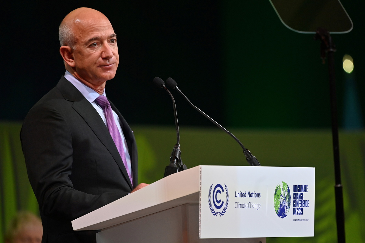 Bezos Earth Fund i CFDA ogłaszają program The Next Thread Initiative. Jeff Bezos zrównoważona moda. Jeff Bezos, w eleganckim ciemnym garniturze, śnieżnobiałej koszuli i purpurowym krawacie, stoi za jasnym podium i wygłasza przemówienie na konferencji. Na mównicy widnieją logotypy i napisy &quot;United Nations Climate Change&quot; oraz &quot;UN CLIMATE CHANGE CONFERENCE UK 2021&quot;. Tło jest zaciemnione, z akcentami zielonego oświetlenia scenicznego.