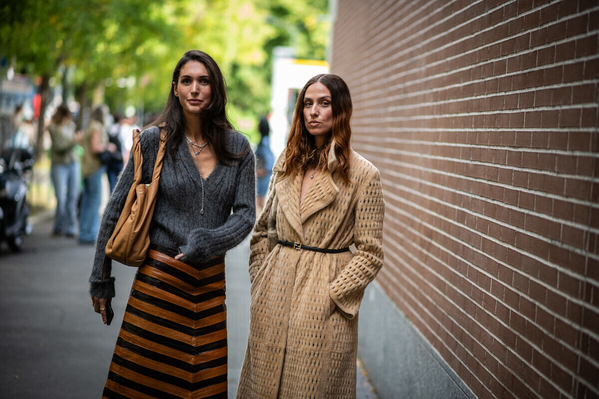 Black Friday 2025 Polska. Dwie kobiety stoją na ulicy obok ściany z brązowej cegły. Kobieta po lewej ma długie ciemne włosy, ubrana jest w szary, zapinany sweter i długą spódnicę w czarne i brązowe, poziome pasy w kształcie jodełki, trzymając brązową skórzaną torbę na ramieniu. Kobieta po prawej ma falowane, brązowe włosy, ma na sobie długi, beżowy płaszcz z dzianiny o fakturowanej powierzchni, przewiązany cienkim czarnym paskiem.