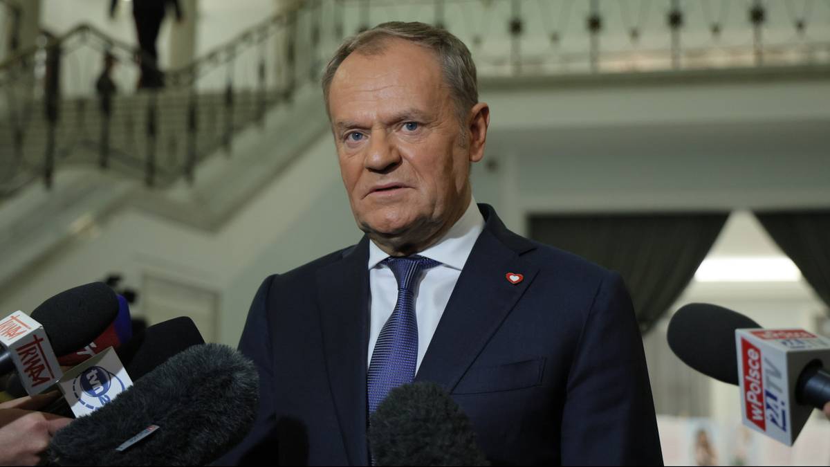 Premier Donald Tusk w garniturze i krawacie stoi przed wieloma mikrofonami, m&oacute;wiąc do dziennikarzy.