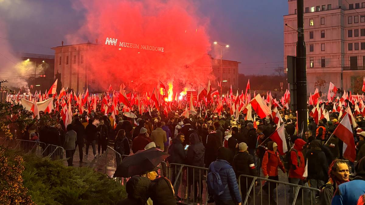 Zgromadzenie ludzi na placu z polskimi flagami, w tle jasnoczerwony dym z rac oświetlający budynki i niebo.