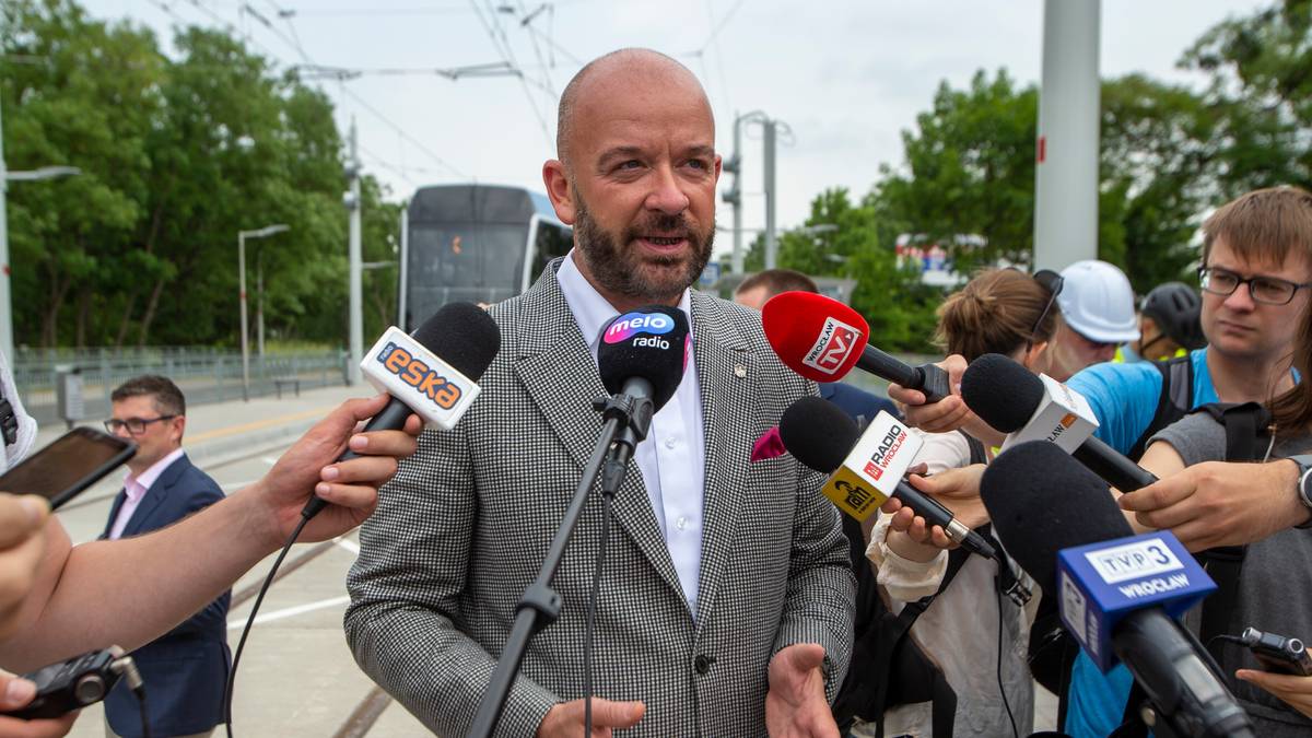 Mężczyzna w jasnej marynarce, otoczony przez dziennikarzy z mikrofonami, przemawia.