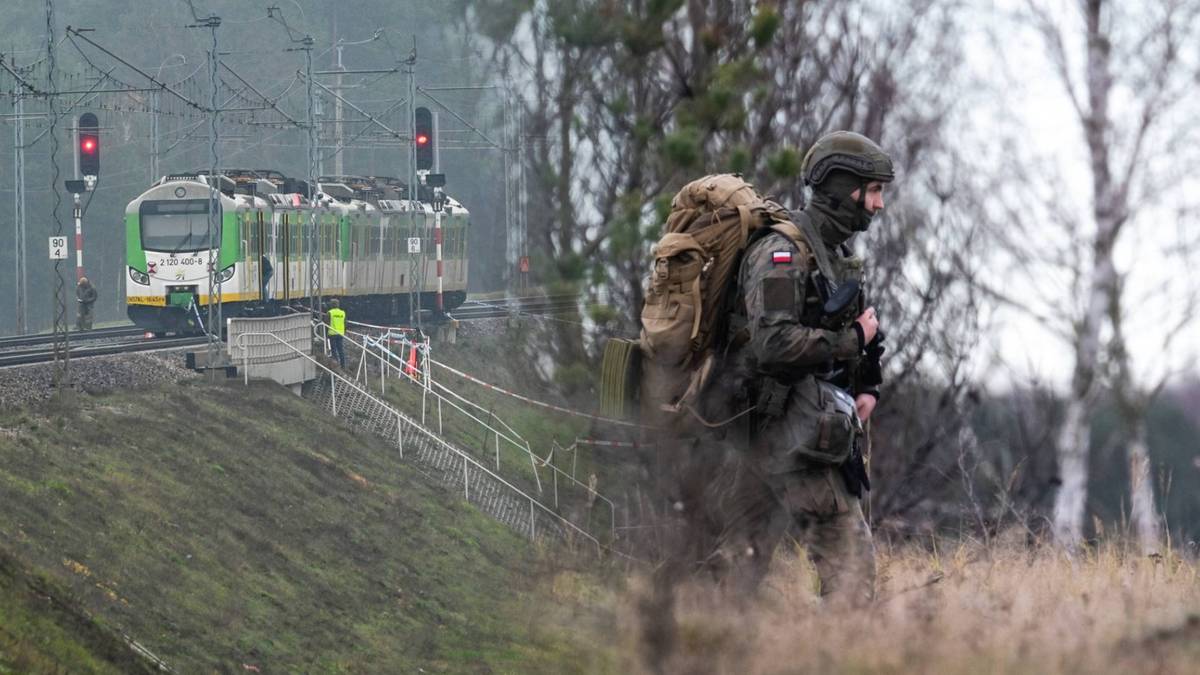 Żołnierz w pełnym umundurowaniu z plecakiem na tle tor&oacute;w kolejowych z pociągiem w tle.