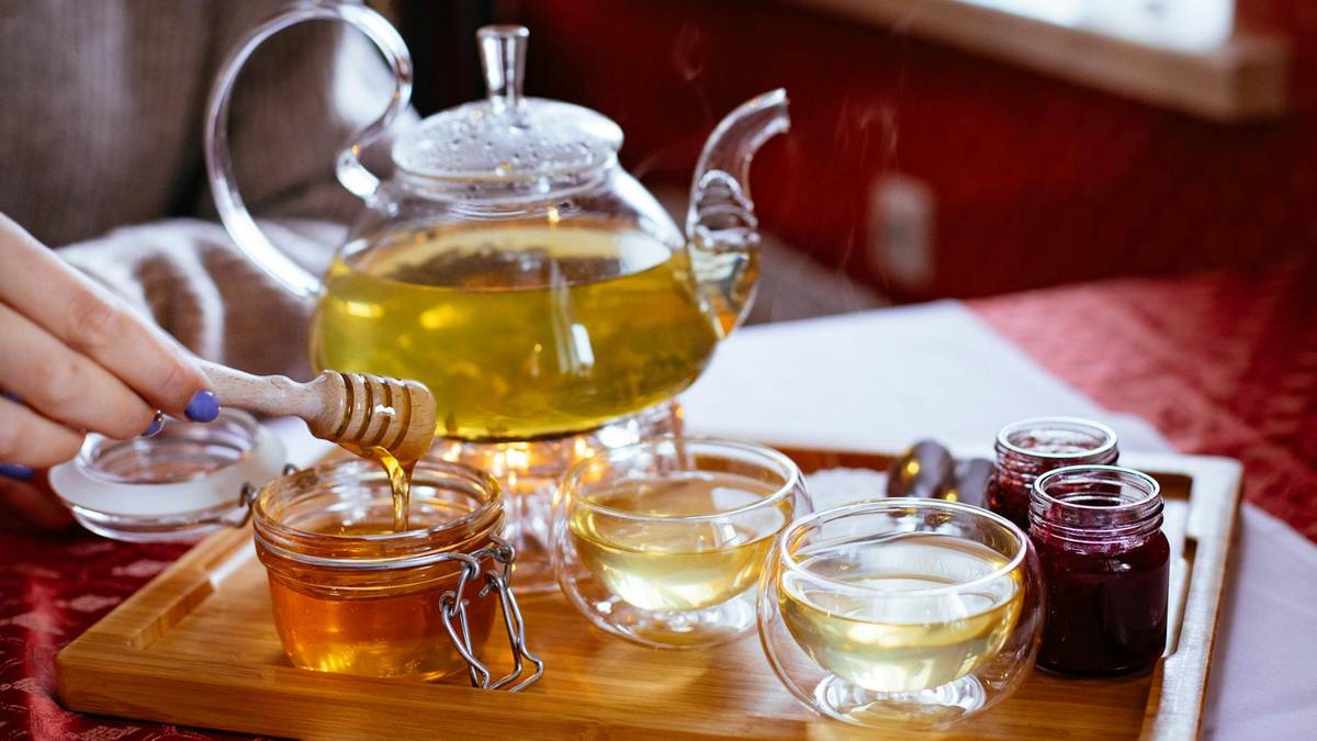 Szklany dzbanek z gorącym napojem, obok dwa kieliszki, słoiczek z miodem i drewniana chochla, osoba dodaje mi&oacute;d do napoju.