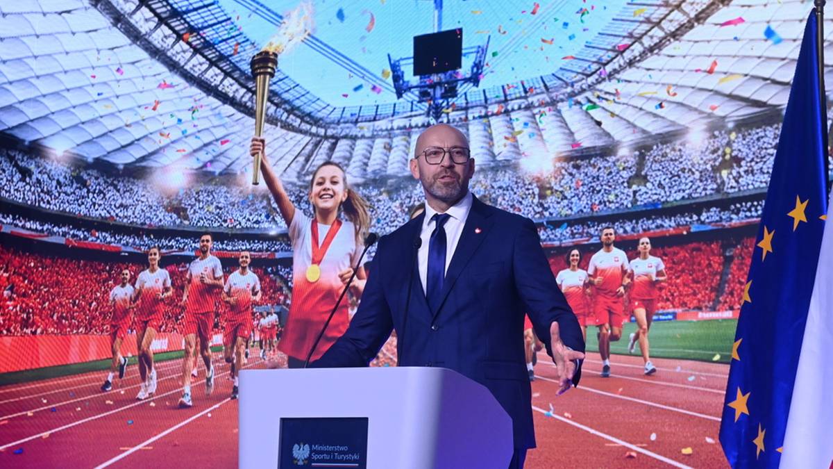 Minister sportu i turystyki Jakub Rutnicki stoi przy m&oacute;wnicy, przemawiając. W tle widoczni są lekkoatleci, flaga olimpijska, flaga Polski i flaga Unii Europejskiej na tle stadionu.