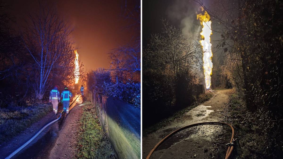 Wysoki słup ognia płonący nocą, z widocznymi sylwetkami strażak&oacute;w i wężem strażackim w pobliżu.