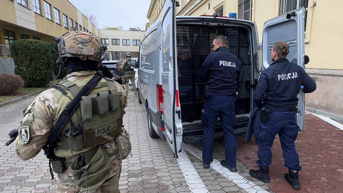 Funkcjonariusze policji w mundurach taktycznych i tradycyjnych otaczają biały van z otwartymi drzwiami, jeden z nich celuje z broni.