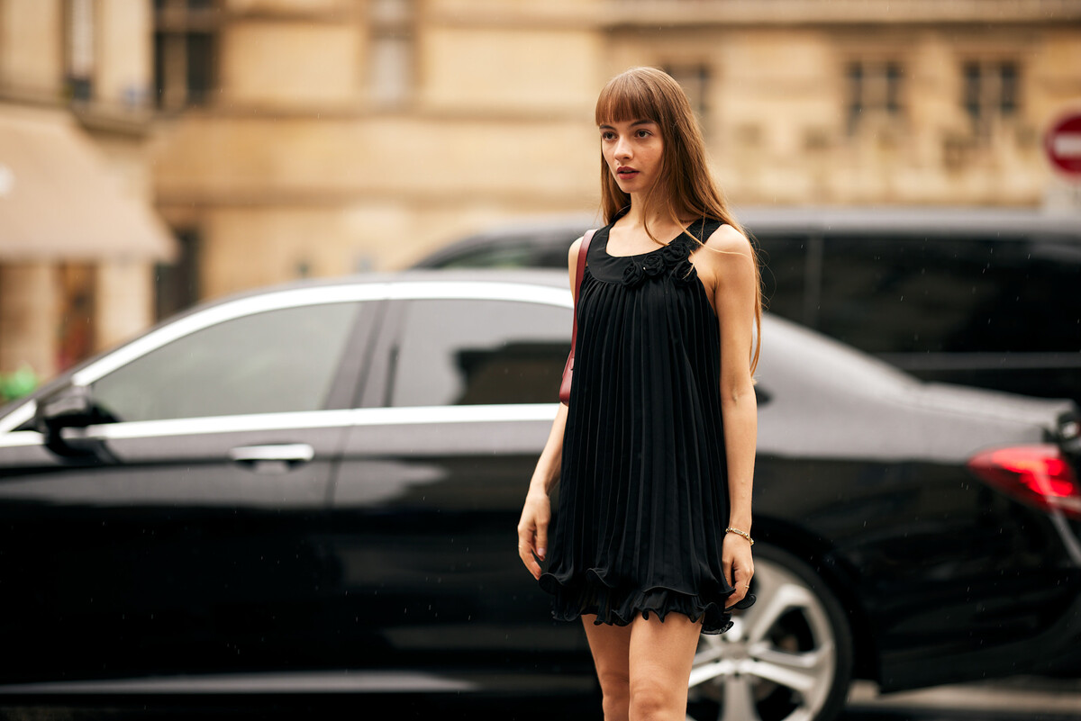 Chosen By klientki, zarzuty. Na zdjęciu: Młoda kobieta w czarnej, plisowanej sukience z odkrytymi ramionami stoi na tle miejskiej ulicy z zaparkowanym samochodem i historyczną architekturą w tle.