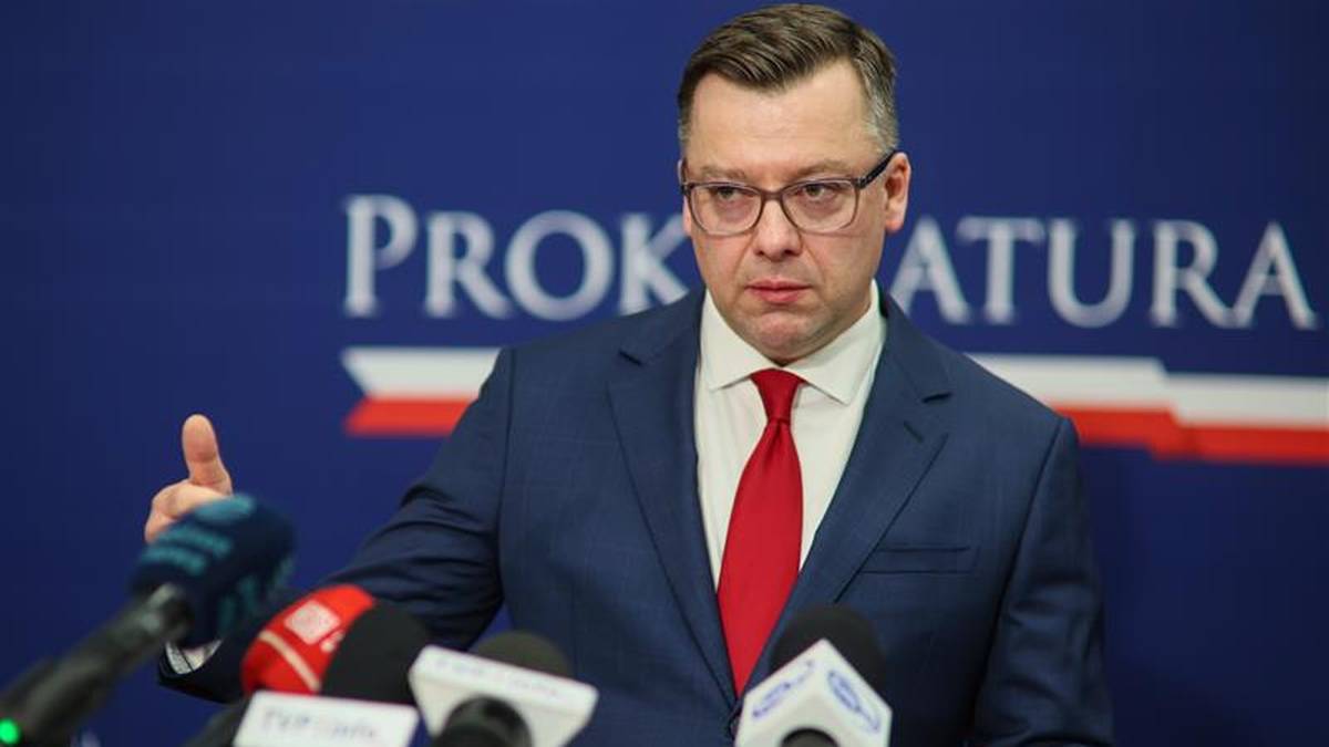 Mężczyzna w garniturze i czerwonym krawacie przemawia przy m&oacute;wnicy, za nim logo prokuratury.