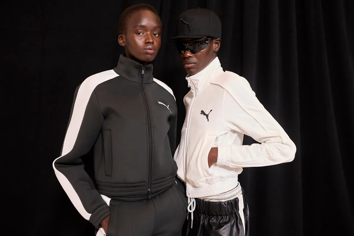 Puma przejęcie przez Anta Sports. Dwie osoby w sportowych strojach Puma – jedna w czerni i bieli, druga w bieli i czerni – prezentują się na tle ciemnej tkaniny.