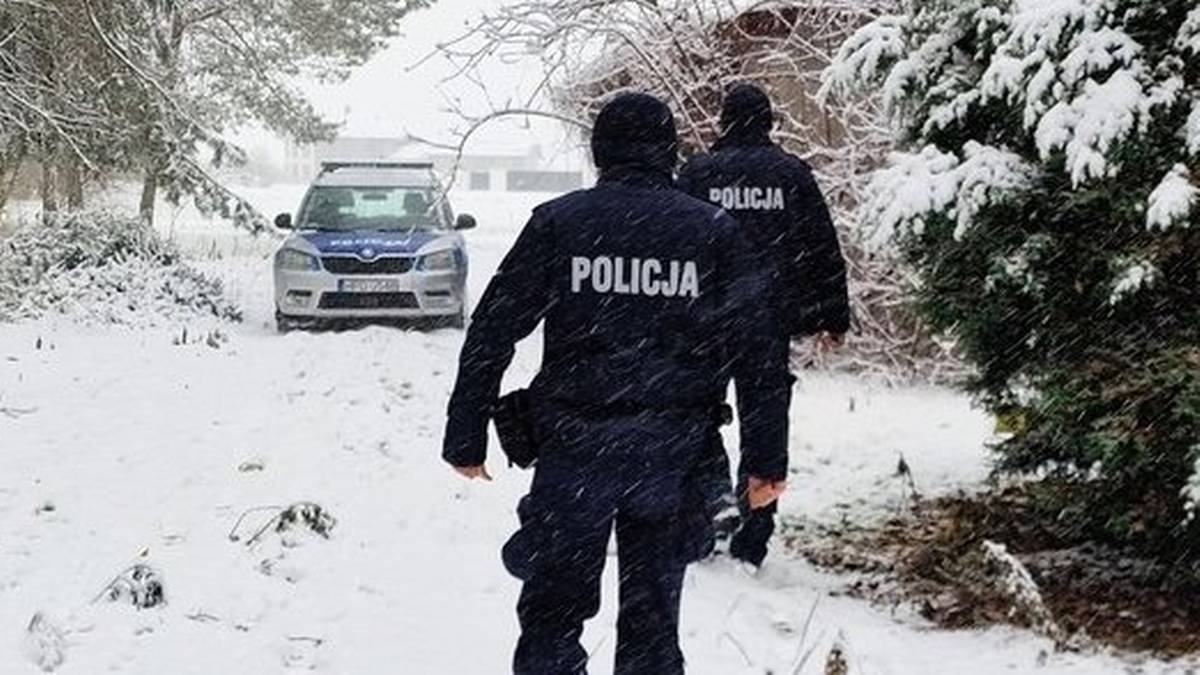 Dw&oacute;ch policjant&oacute;w w mundurach idzie przez zaśnieżony teren leśny w kierunku samochodu policyjnego.