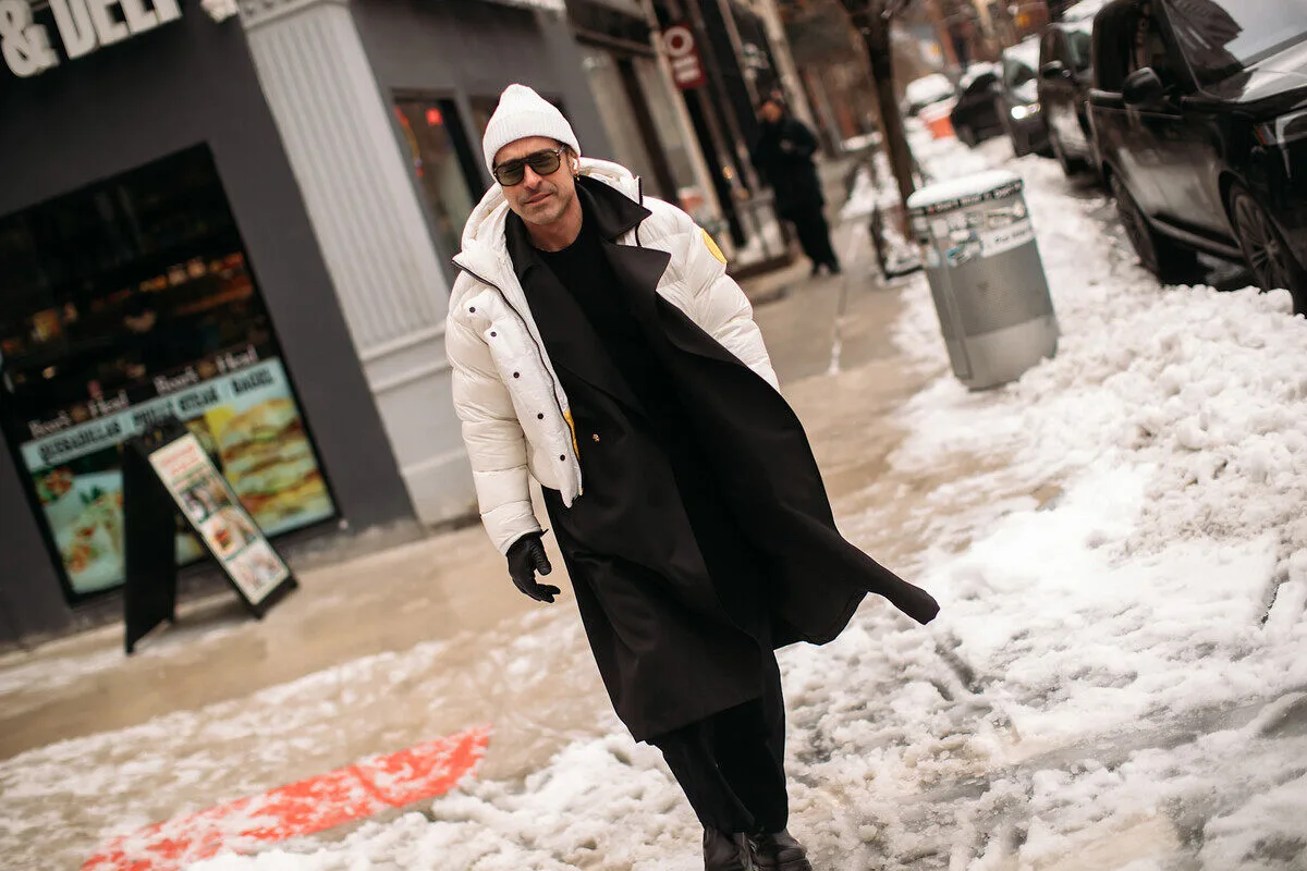 PFAS kurtki C&amp;A. Mężczyzna odziany w biały puchowy okrycie wierzchnie, czarny długi płaszcz, białą czapkę i ciemne okulary przemierza chodnik pokryty śniegiem w miejskiej scenerii. Ulica prezentuje zimowy krajobraz, z zaparkowanymi autami, błotem pośniegowym i sklepami w tle. Postać prezentuje się szykownie i pewnie, zmierzając w stronę obiektywu.