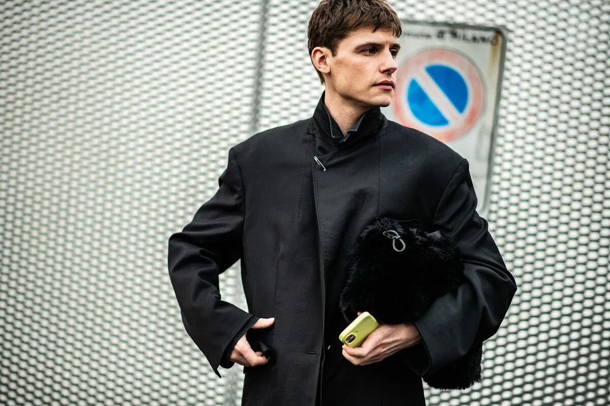 Greenwashing UOKiK Tchibo, Bolt. Portret mężczyzny w czarnym, minimalistycznym płaszczu ze stójką, spiętym agrafką pod szyją. Mężczyzna trzyma pod pachą puszystą, czarną torbę oraz żółty telefon, stojąc na tle metalowej siatki i znaku drogowego.