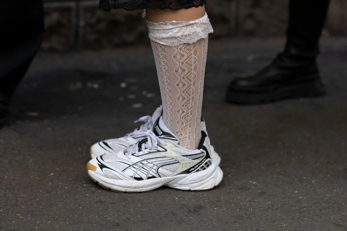 Zbliżenie na nogi kobiety w białych koronkowych podkolanówkach i sportowych butach Puma Velophasis. Kreacja łączy w sobie pierwiastki romantyzmu z duchem nurtu dad shoes, w czasie gdy w tle zmian branżowych dystrybutor obuwia sportowego likwiduje punkty stacjonarne na rzecz handlu w sieci.