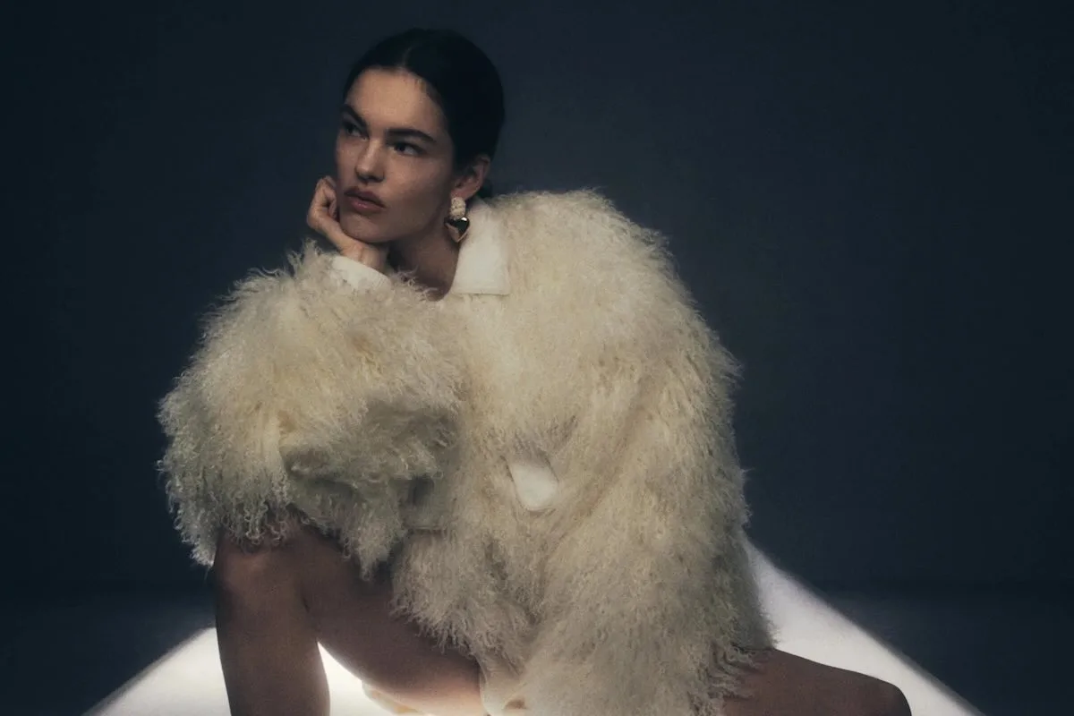 Modelka w jasnym, futrzanym okryciu o formie architektonicznej, siedząca w minimalistycznym studiu, prezentująca stylistykę nowoczesnego damskiego szycia marki premium.