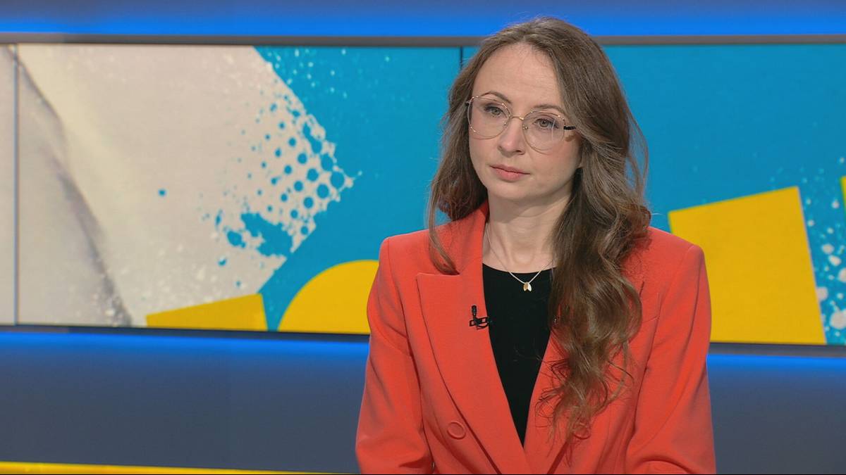 Kobieta w okularach i pomarańczowej marynarce siedzi przed ekranem telewizyjnym z kolorowym wzorem.