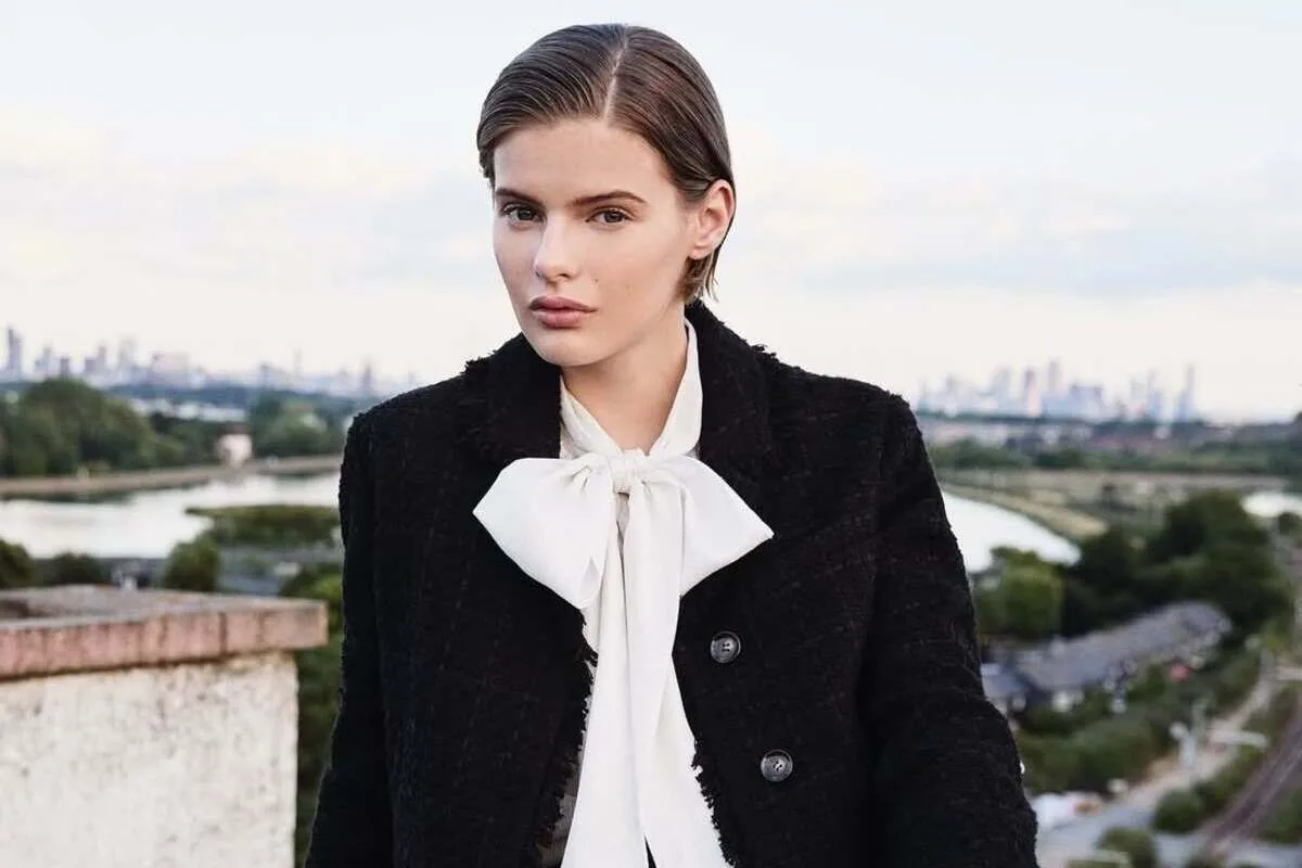 LK Bennett zamyka sklepy. Młoda kobieta w czarnym płaszczu i białej bluzce z wiązaną kokardą stoi na tle rzeki i panoramy miasta, patrząc prosto w obiektyw.