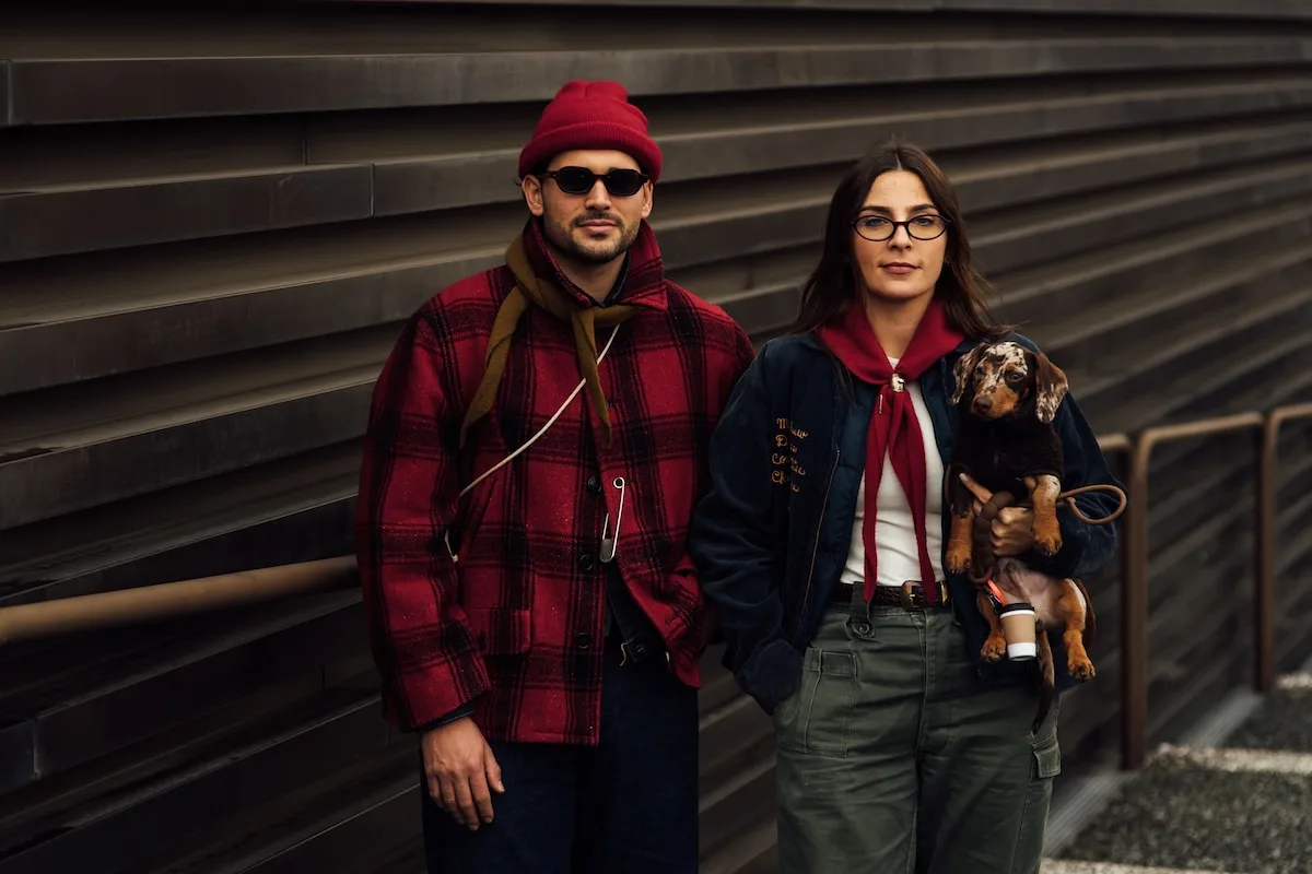 Niedziele handlowe 2026. Czy 18 stycznia są otwarte sklepy? Na zdjęciu para. Dwoje stylowo ubranych dorosłych stojących przy nowoczesnej, metalowej ścianie; mężczyzna w czerwonej czapce i kraciastej kurtce, kobieta w okularach trzymająca jamnika, miejski look w jesienno-zimowym stylu.
