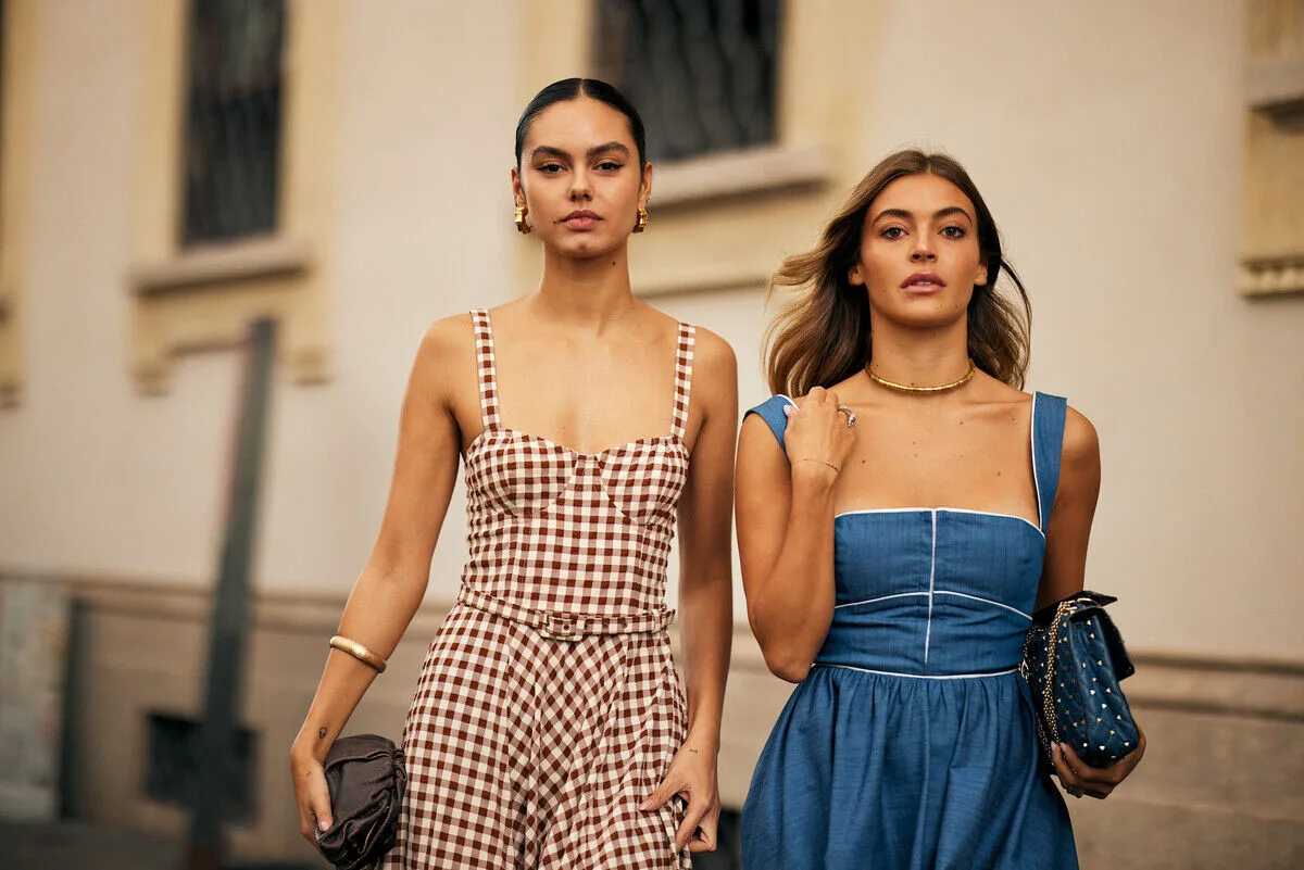Najlepsze luksusowe platformy resale 2026. Dwie młode kobiety spacerują po ulicy w krajobrazie miejskim, mają na sobie letnie kreacje — jedna w brązowo-białą kratę, druga w błękitną — trzymają eleganckie torebki i emanują pewnością siebie oraz stylem.