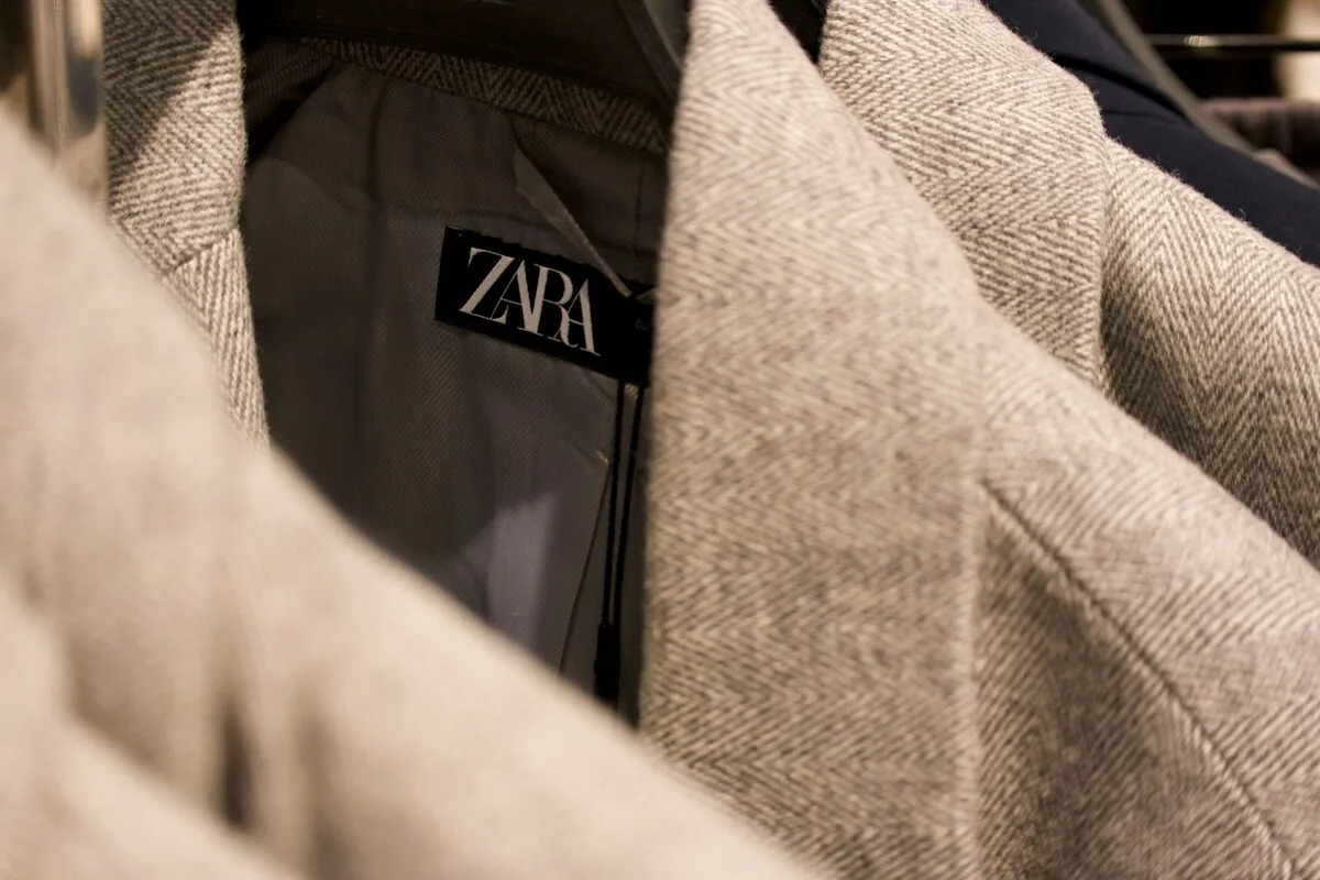 Greenwashing Zara UOKiK. Detal odzieży w sklepie: czarna metka marki ZARA widoczna na tle podszewki szarego płaszcza lub marynarki. Materiał wierzchni ma wyraźny splot w jodełkę (herringbone). W tle widoczne inne ubrania na wieszakach.