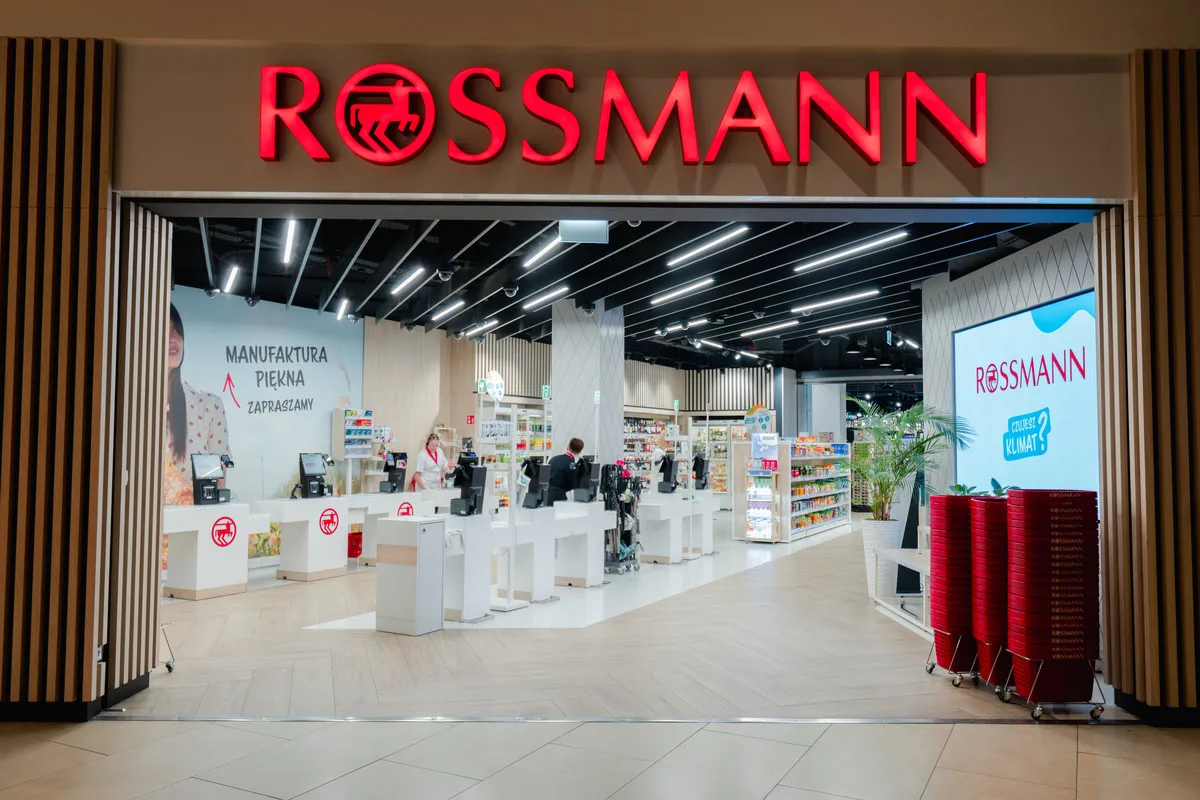Rossmann apteka internetowa. Ukazanie wejścia do współczesnej drogerii z szyldem Rossmann, kasami samoobsługowymi, półkami z produktami oraz czerwonymi koszykami na zakupy.