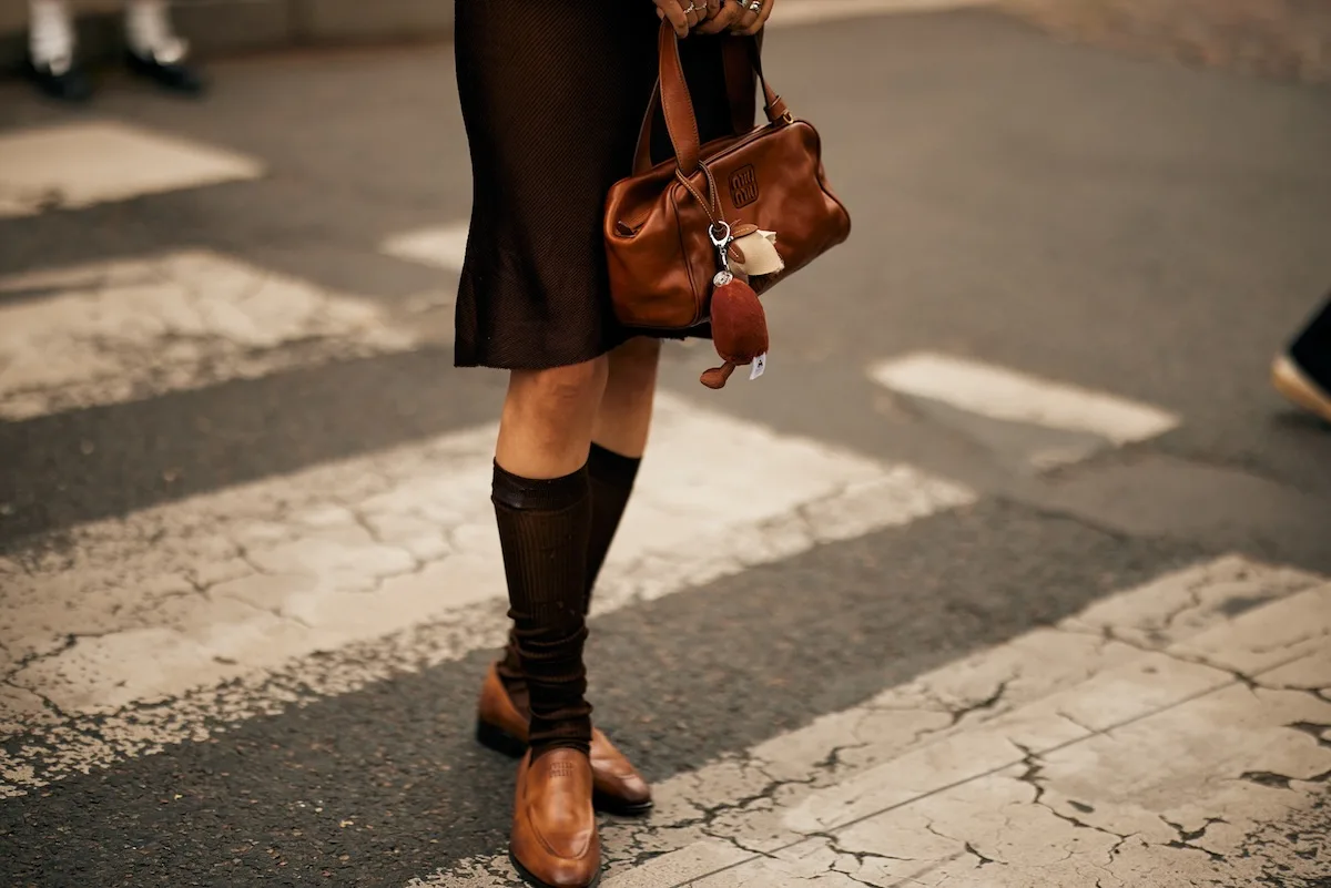 167 zwolnień w De Bijenkorf. Luksusowy gigant reorganizuje biznes. Na zdjęciu zbliżenie na nogi modelki trzymające w rękach brązową torebkę.