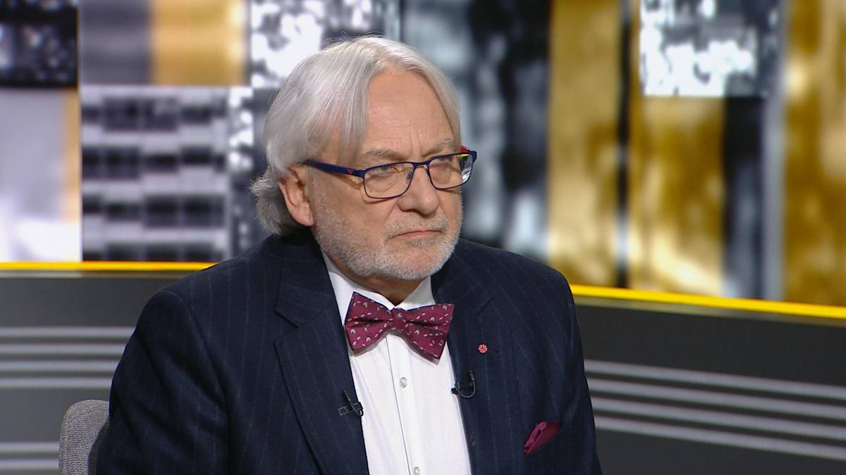 Profesor Wojciech Maksymowicz w okularach, ubrany w garnitur z muszką, siedzi w studiu telewizyjnym.