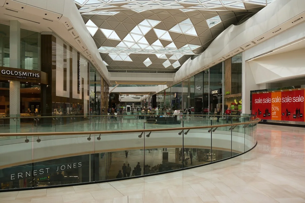 Przebudowa Alfa Centrum w Gdańsku. Wnętrze nowoczesnej galerii handlowej z przeszklonym dachem, ciągami sklepów i widocznymi punktami sprzedaży po obu stronach, w tle klienci robiący zakupy.