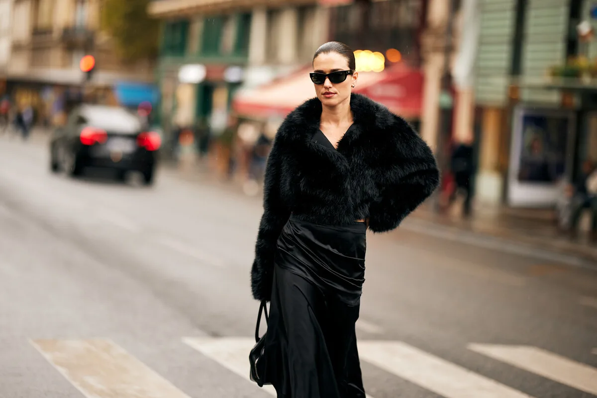 Długi Saks Global. Dama w szykownej, czarnej kreacji przechodzi przez przejście dla pieszych na ulicy miasta. Nosi krótkie, puszyste okrycie wierzchnie, atłasową spódnicę i ciemne okulary słoneczne, a jej włosy są elegancko uczesane. W tle widać zamazany ruch uliczny, budowle oraz wystawy sklepowe, co nadaje całości metropolitalny charakter.