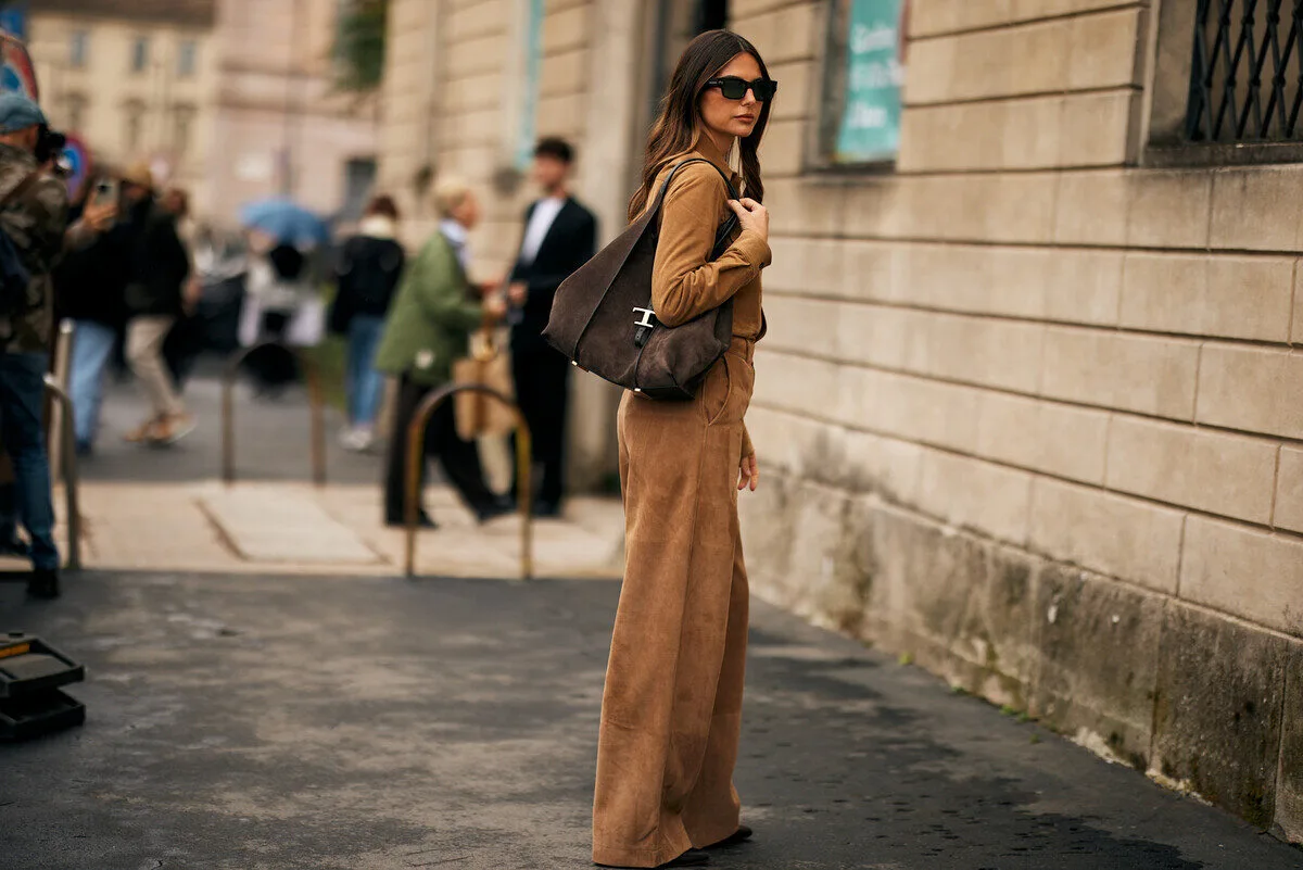 Zalando i Temu ukarane przez UOKiK. Elegancka kobieta stoi na miejskim chodniku przy kamiennej ścianie, ubrana w brązowy, zamszowy komplet i ciemne okulary przeciwsłoneczne. Na ramieniu ma dużą brązową torbę, a w tle widać rozmytych przechodniów i zabudowę miejską, co nadaje zdjęciu modowy, streetstyle’owy charakter.