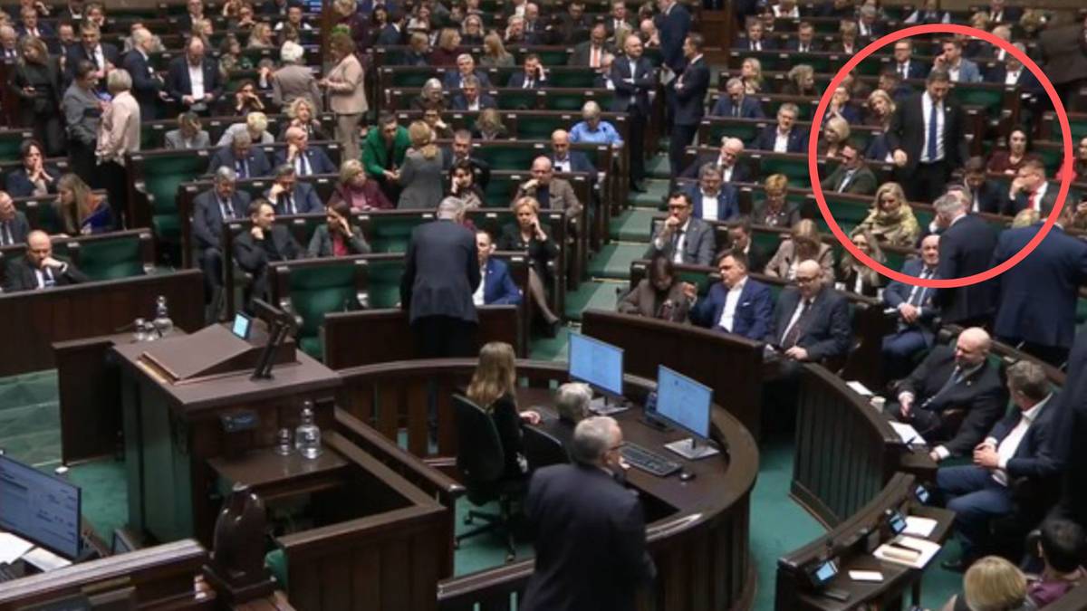 Przemawiający mężczyzna w garniturze stoi przed grupą siedzących posł&oacute;w w dużej sali parlamentarnej.